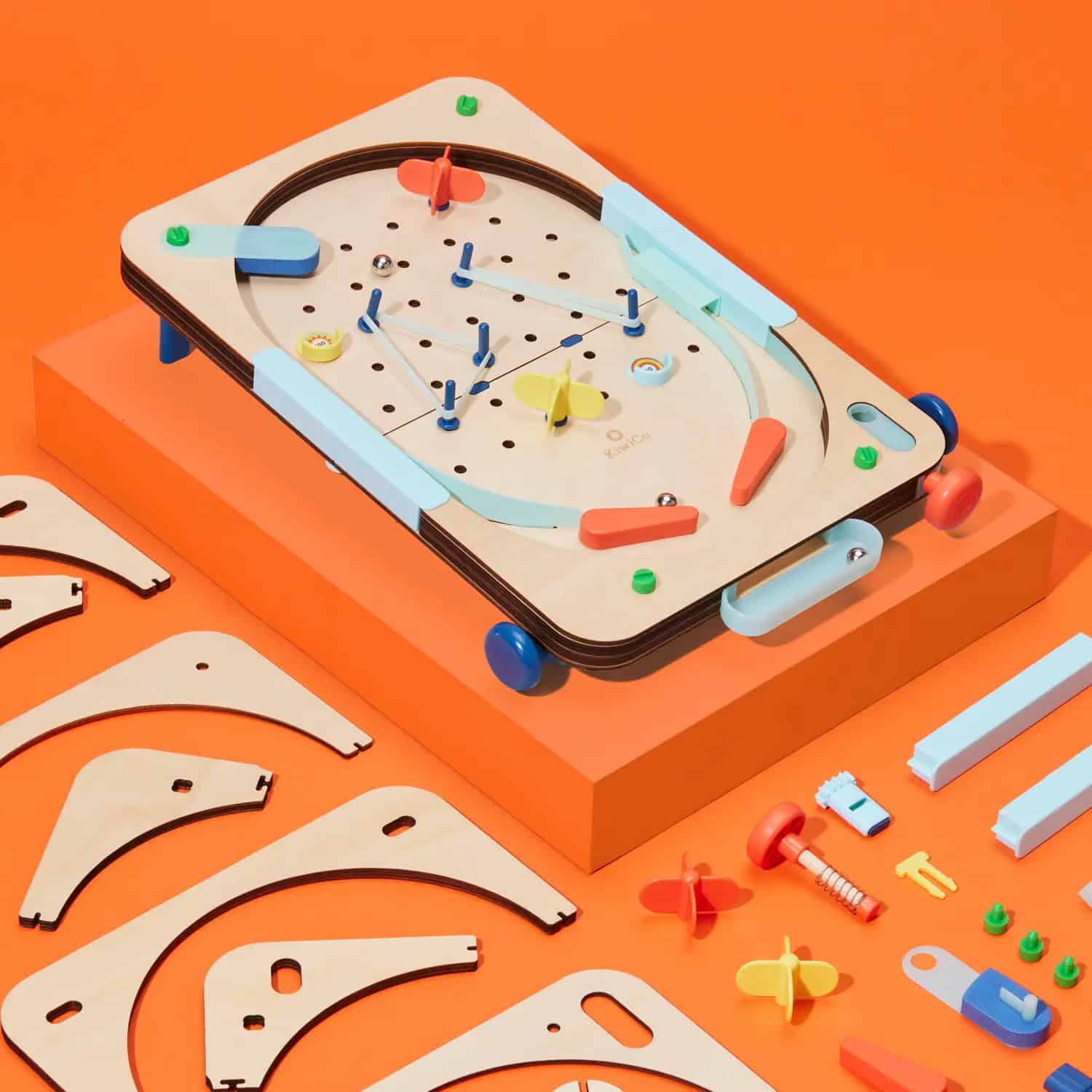 A wooden pinball game set with colorful components is displayed on an orange platform, surrounded by unassembled wooden and plastic parts—an engaging way to help prevent summer learning loss against a vibrant orange background.