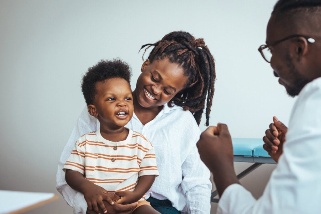 A child sits on a womans lap, both smiling, while they interact with a person in a white coat—perhaps discussing 2-year-old milestones like how many words should a 2-year-old know or how much sleep does a 2-year-old need.