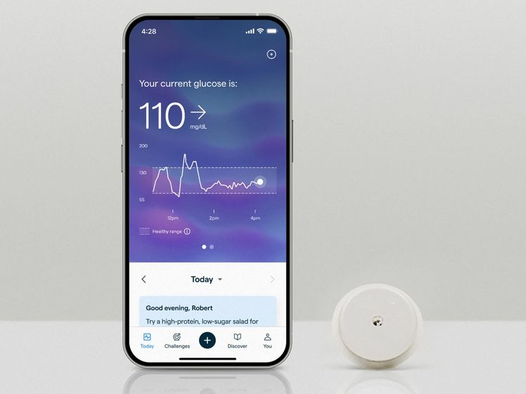 Abbott Lingo Continuous Blood Glucose Monitor, a small white disc, beside a mobile phone showing the complimentary app that shows glucose score