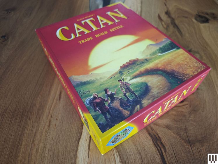 Box for a boardgame on wooden surface. Front says Catan and is a scene of travelers looking down a long road and a town in the sunset distance.