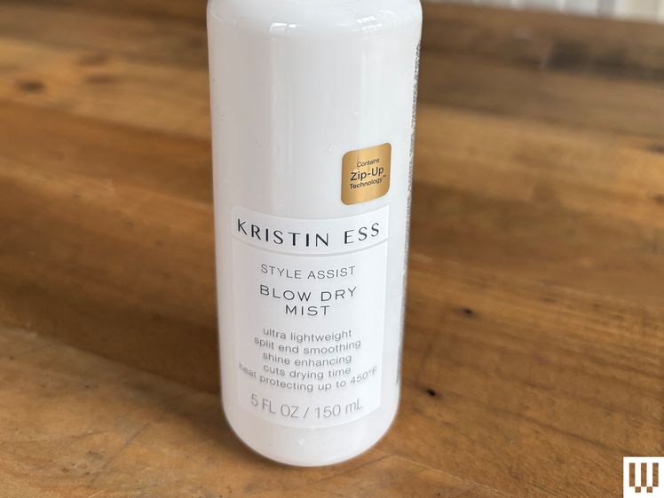 Kristin Ess Style Assist Blow-Dry Mist, a white bottle with spray top, sitting on a wooden table