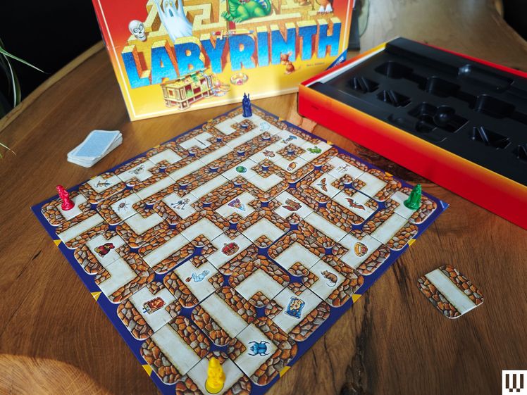 Contents of the Labyrinth board game on a wooden surface. In the back is the game box, center is the board with puzzle-like pathways and small plastic pieces, and to the right is the empty packaging.