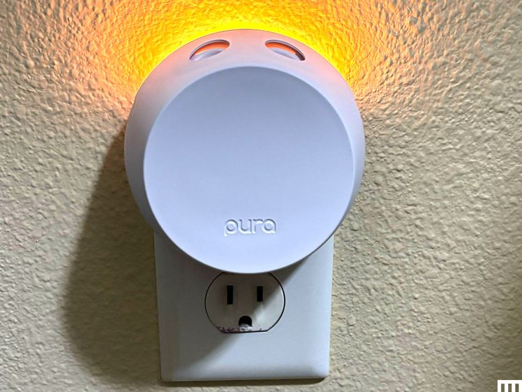 Pura Smart Diffuser, a white disc-shaped device with two holes in the top plugged into an outlet while casting a soft yellow light from the top