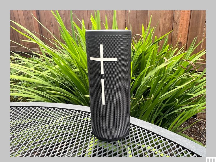Front view of the Ultimate Ears Boom 4, a black cylindrical speaker with a large plus and minus sign, sitting on a metal outdoor table